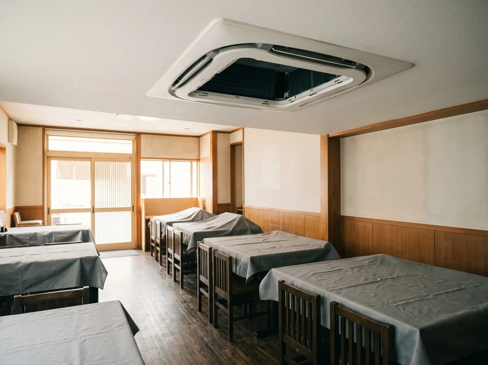 営業前の飲食店の客席で、テーブルと椅子がシートで覆われ、天井のカセット形エアコンの吸込グリルが外されている様子